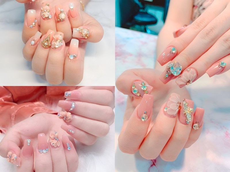 Bàn tay với móng tay đính đá quý và hoa nổi, thiết kế tinh xảo trên nền màu hồng nhạt.