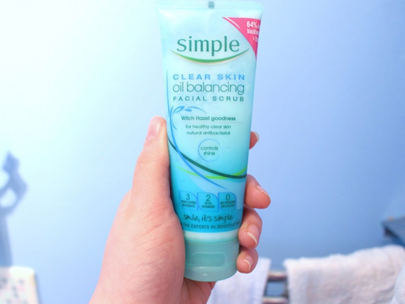 Một bàn tay đang cầm tuýp sữa rửa mặt màu xanh, nhãn hiệu "Simple", với nền màu xanh dương nhạt.