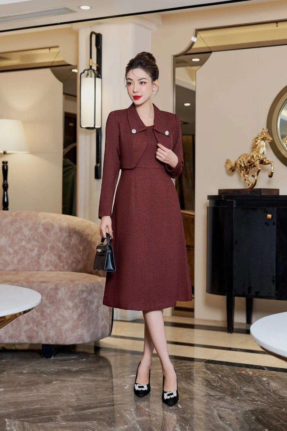 Người phụ nữ mặc váy đỏ đứng trong phòng khách sang trọng, tay cầm túi nhỏ, nền có ghế sofa và tượng ngựa.