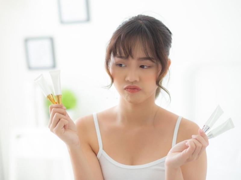 4d5b435c-kem-lot-cho-da-kho-2 Người phụ nữ cầm nhiều tuýp mỹ phẩm, vẻ mặt lưỡng lự, ngồi trong phòng sáng sủa.
