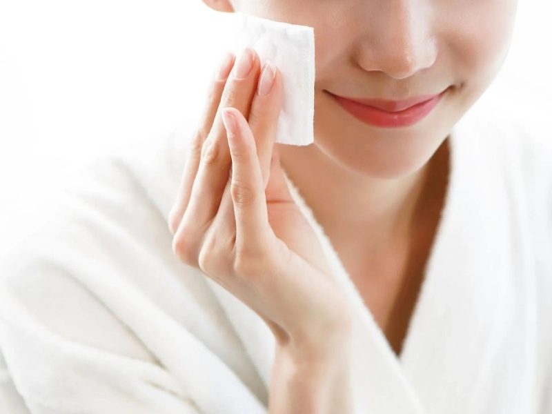 Người mặc áo choàng trắng đang dùng bông cotton lau má trên khuôn mặt, tạo cảm giác thư giãn và chăm sóc da.