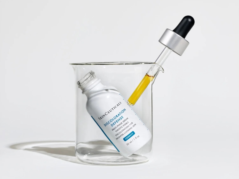 Chai serum trắng và ống nhỏ giọt vàng trong cốc thủy tinh trên nền trắng.