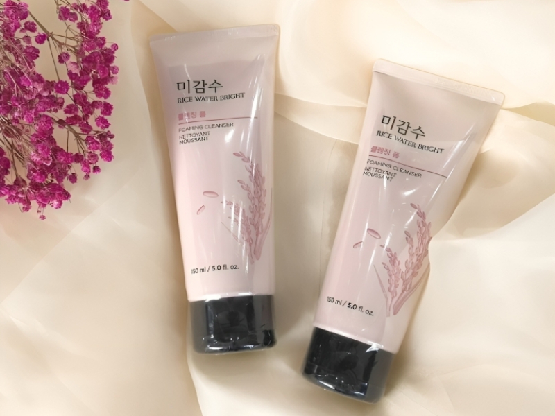Hai tuýp sữa rửa mặt trên nền vải màu kem, cạnh đó là cành hoa màu hồng.