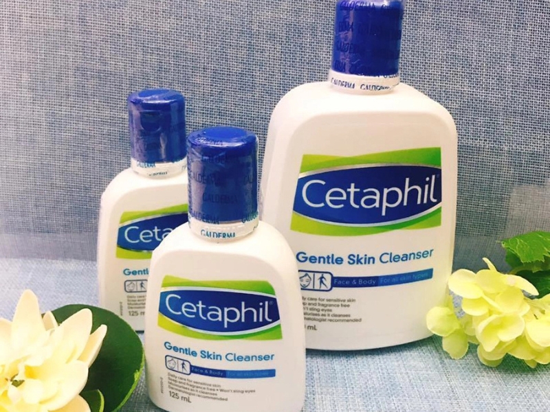 63f395fd-sua-rua-mat-cho-da-hon-hop-thien-kho-03 Ba chai sữa rửa mặt nhãn hiệu Cetaphil với nền vải xanh và hoa trang trí bên cạnh.