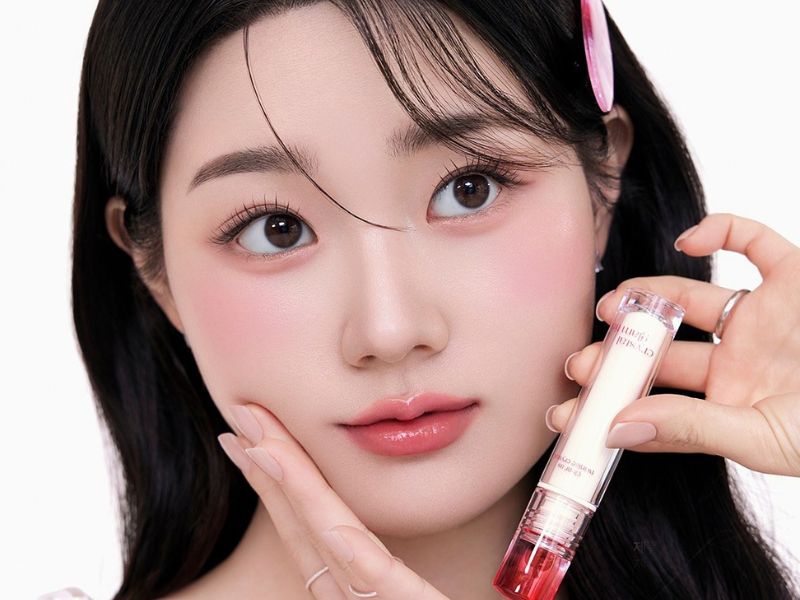 643f32ae-son-mau-hong-dat-dep-17 Người cầm thỏi son dưỡng môi, tay chạm má, tóc cài kẹp, trang điểm nhẹ nhàng, nền trắng.