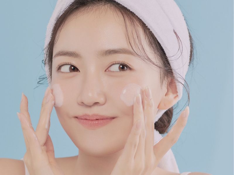 Người đeo băng đô trắng, thoa kem dưỡng da lên má với nền xanh dương.