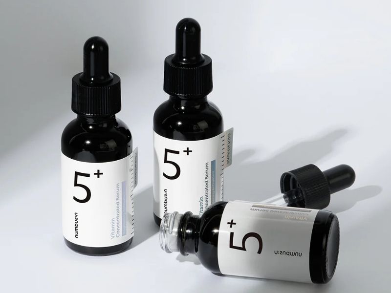 6f07dc7e-serum-lam-deu-mau-da-13 Ba chai serum màu đen có nhãn 5+, một chai nghiêng với nắp mở và ống nhỏ giọt bên cạnh.