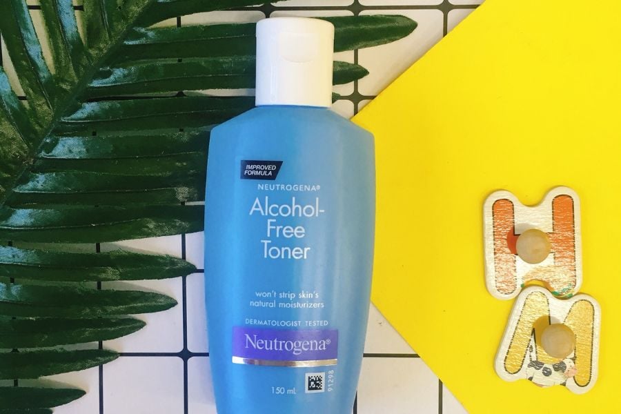 Chai toner màu xanh, không chứa cồn, đặt trên nền gạch trắng với lá cây và vật dụng trang trí màu vàng.
