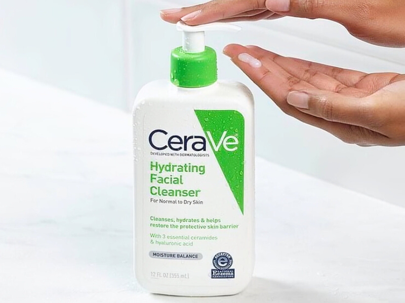 771531cb-sua-rua-mat-cho-da-kho-nhay-cam-6 Chai sữa rửa mặt dưỡng ẩm CeraVe, nắp màu xanh lá, tay đang ấn lấy sản phẩm, nền trắng.