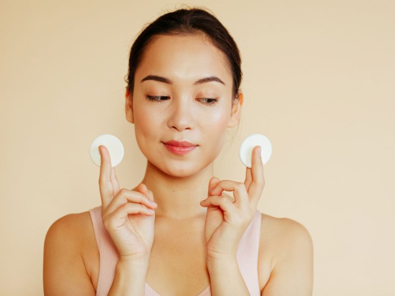 78cf786b-nuoc-tay-trang-tot-nhat-hien-nay-25 Người phụ nữ cầm hai bông tẩy trang, đứng trên nền màu be, mặc áo không tay màu hồng nhạt.