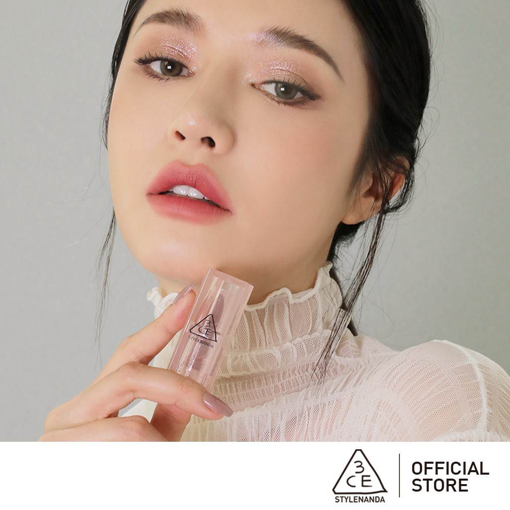 80fae968-xx-compressed-1 Người mẫu trang điểm tinh tế, cầm thỏi son 3CE, mặc áo ren trắng, nền xám, logo "OFFICIAL STORE" bên dưới.