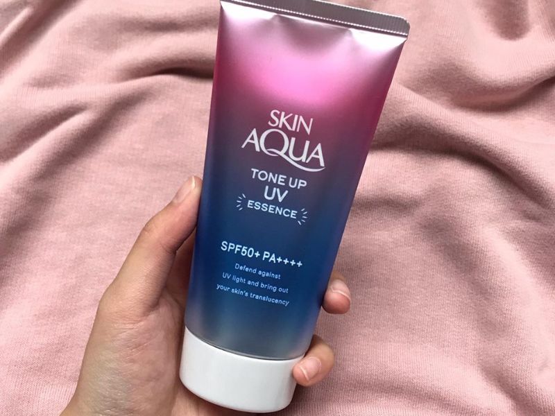 83d70ad2-review-kem-chong-nang-skin-aqua14 Tay cầm tuýp kem chống nắng Skin Aqua Tone Up UV Essence trên nền vải màu hồng nhạt.