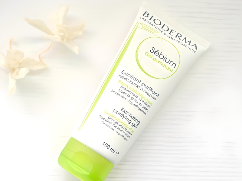 84ce48e5-bione Tuýp gel tẩy tế bào chết thương hiệu Bioderma nằm trên nền trắng, bên cạnh có hai bông hoa khô trang trí.
