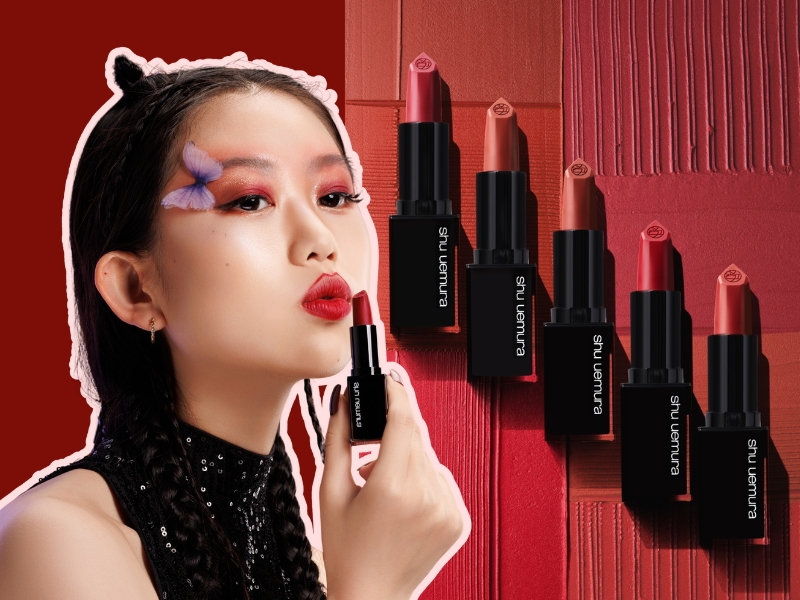 89922257-review-son-shu-uemura-3 Người phụ nữ trang điểm môi đỏ, cầm thỏi son, nền là nhiều thỏi son khác nhau trên bề mặt màu đỏ.