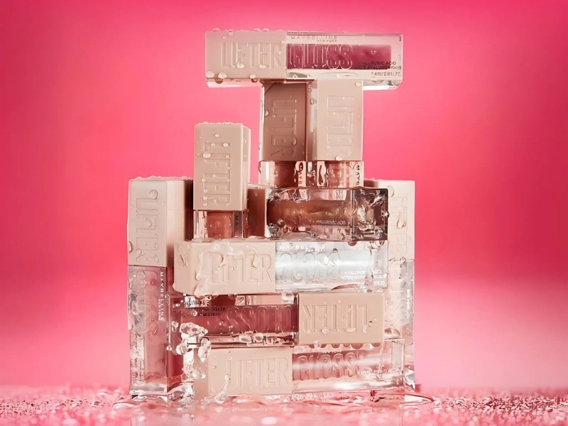 92cd77cd-review-son-maybelline-2 Các chai mỹ phẩm xếp chồng trên nền hồng, có giọt nước đọng trên bề mặt.