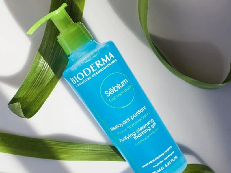 9437ce71-sua-rua-mat-cho-da-dau-01 Chai gel rửa mặt màu xanh có vòi bơm, nhãn hiệu Bioderma, đặt trên nền trắng với lá trang trí.
