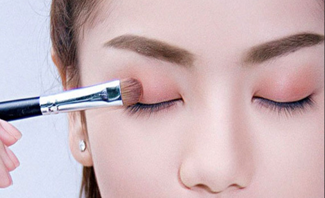 Cận cảnh một người đang sử dụng cọ trang điểm tán phấn mắt màu hồng nhạt lên vùng mí mắt.