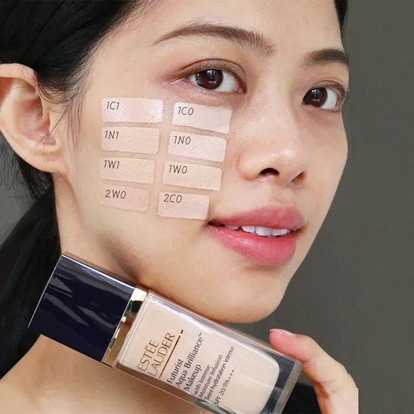 Người cầm chai kem nền, trên má có các mẫu thử màu kem với mã số khác nhau.