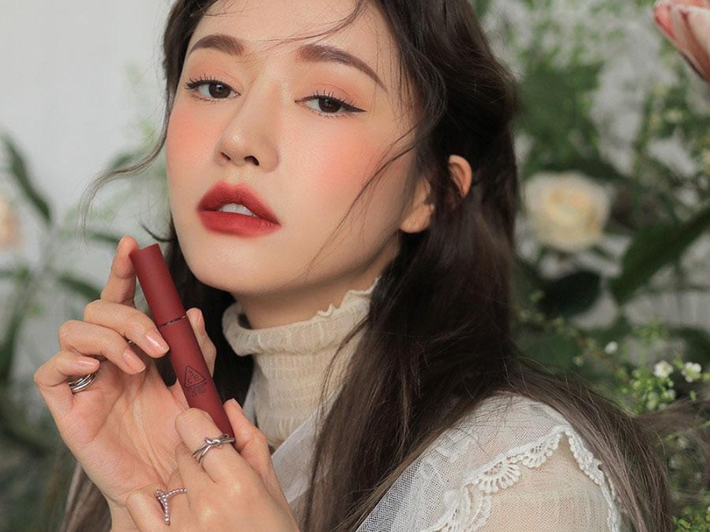 a0d7bd73-son-mau-do-gach-dep-8 Người phụ nữ cầm son môi đỏ, trang điểm tinh tế, nền là cây cỏ và hoa hồng trắng.