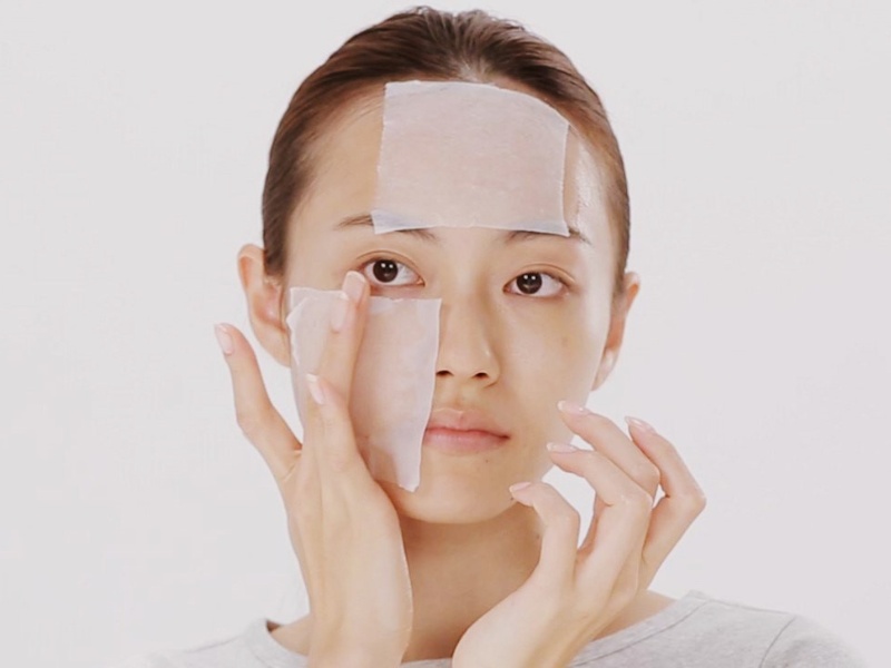 Người phụ nữ đang đắp mặt nạ giấy dưỡng da trên trán và má, nền trắng.