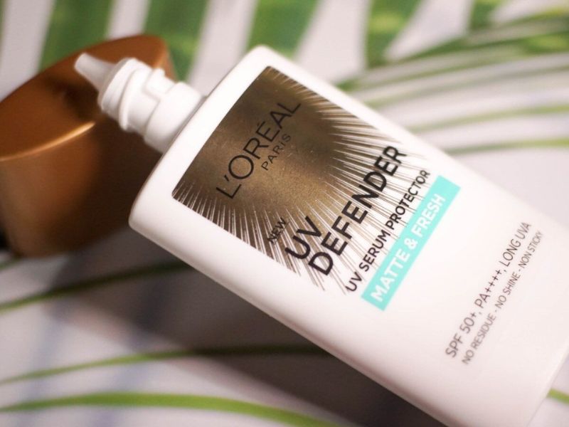 aac55381-kem-chong-nang-duong-am-13 Chai serum chống nắng L'Oreal UV Defender với nắp vàng, nằm trên nền lá xanh.