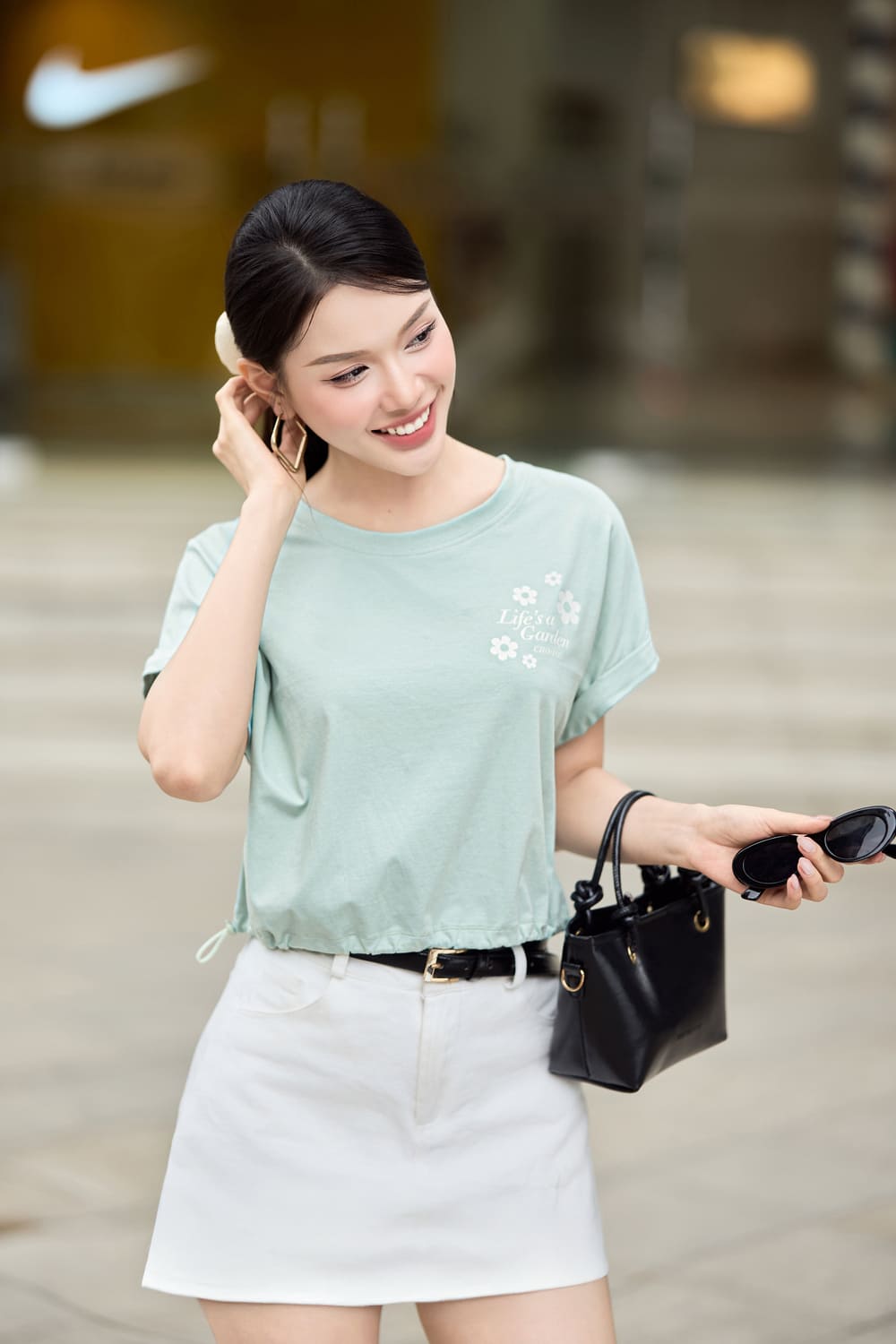 Người phụ nữ mặc áo thun xanh nhạt và váy trắng, cầm kính mát và túi xách đen, đứng trên phố.