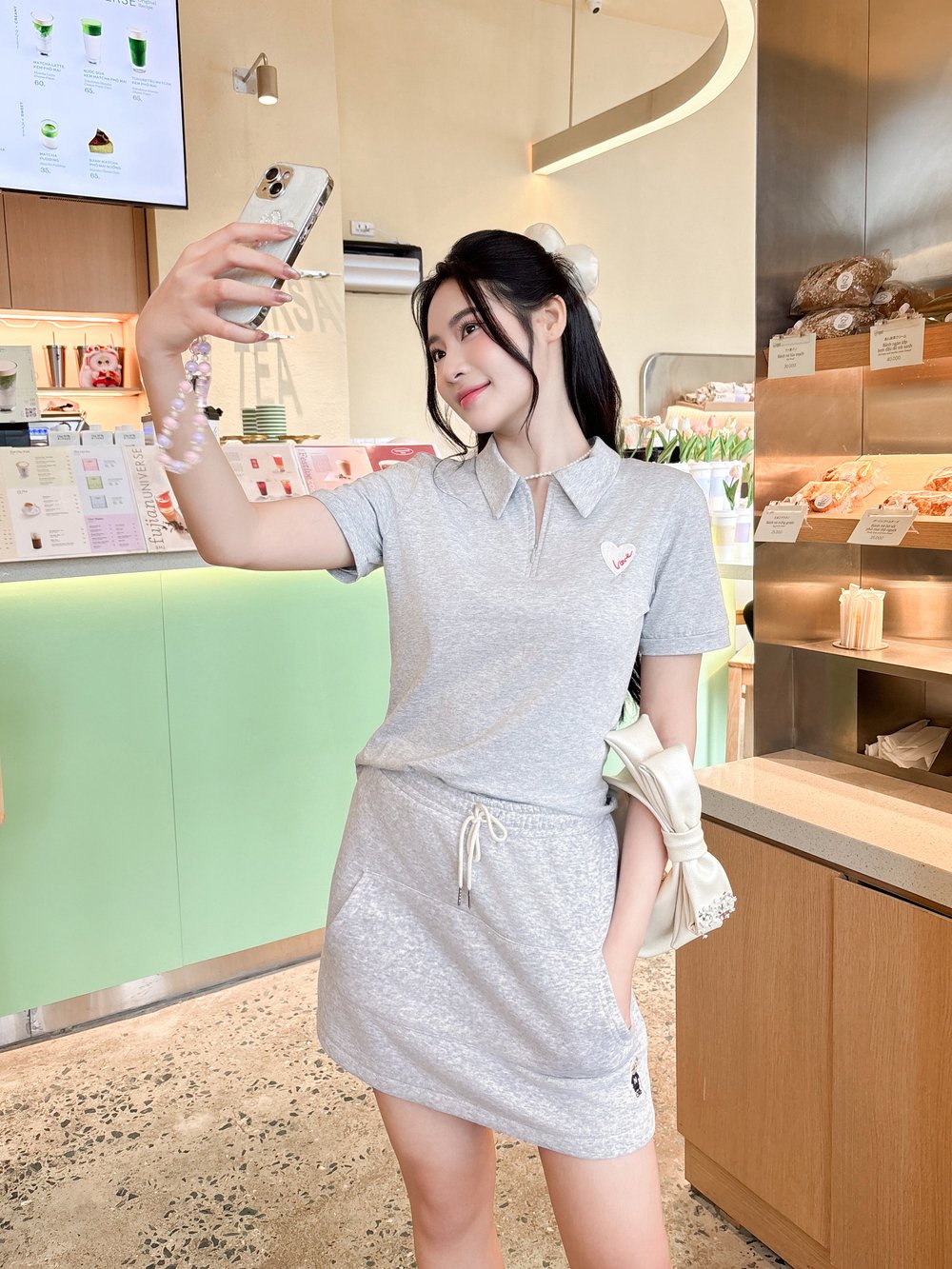 Người phụ nữ mặc váy xám chụp ảnh selfie trong quán cà phê với nền menu và bánh ngọt.
