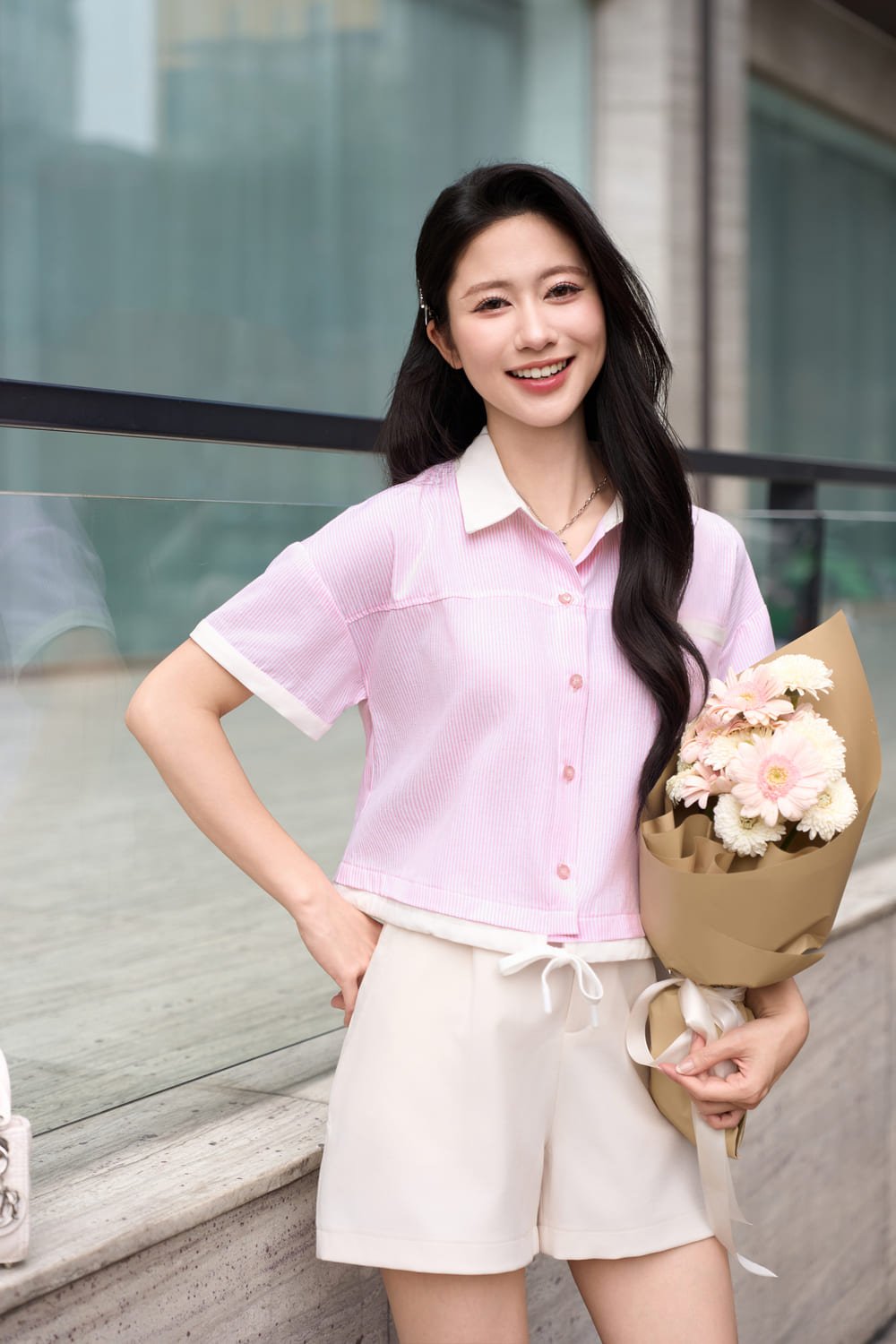 Người mặc áo sơ mi hồng, quần trắng, cầm bó hoa, đứng cạnh bức tường kính, mỉm cười nhìn vào máy ảnh.