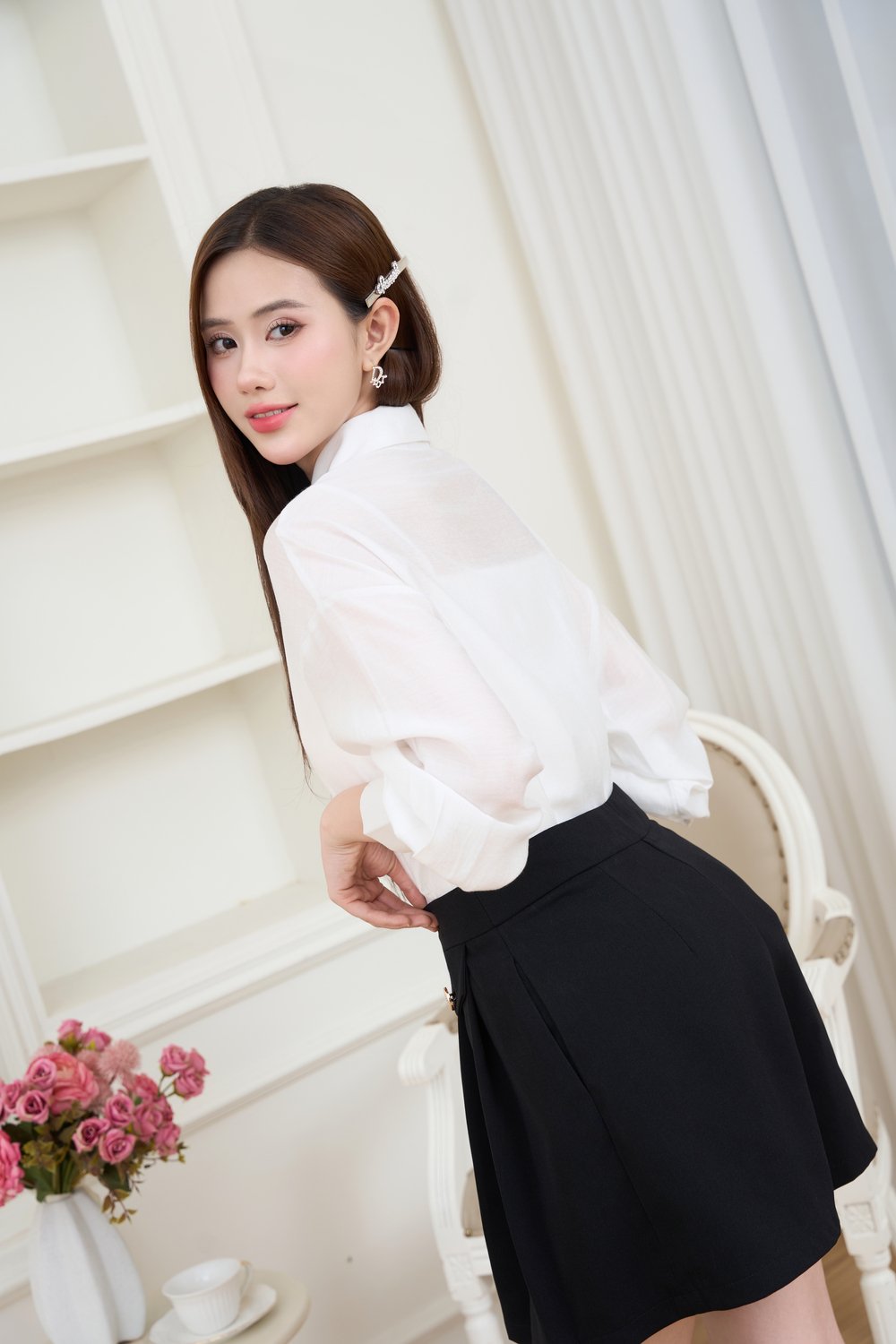 Người mặc áo sơ mi trắng và váy đen đứng cạnh bàn có bình hoa hồng trong phòng sáng sủa.