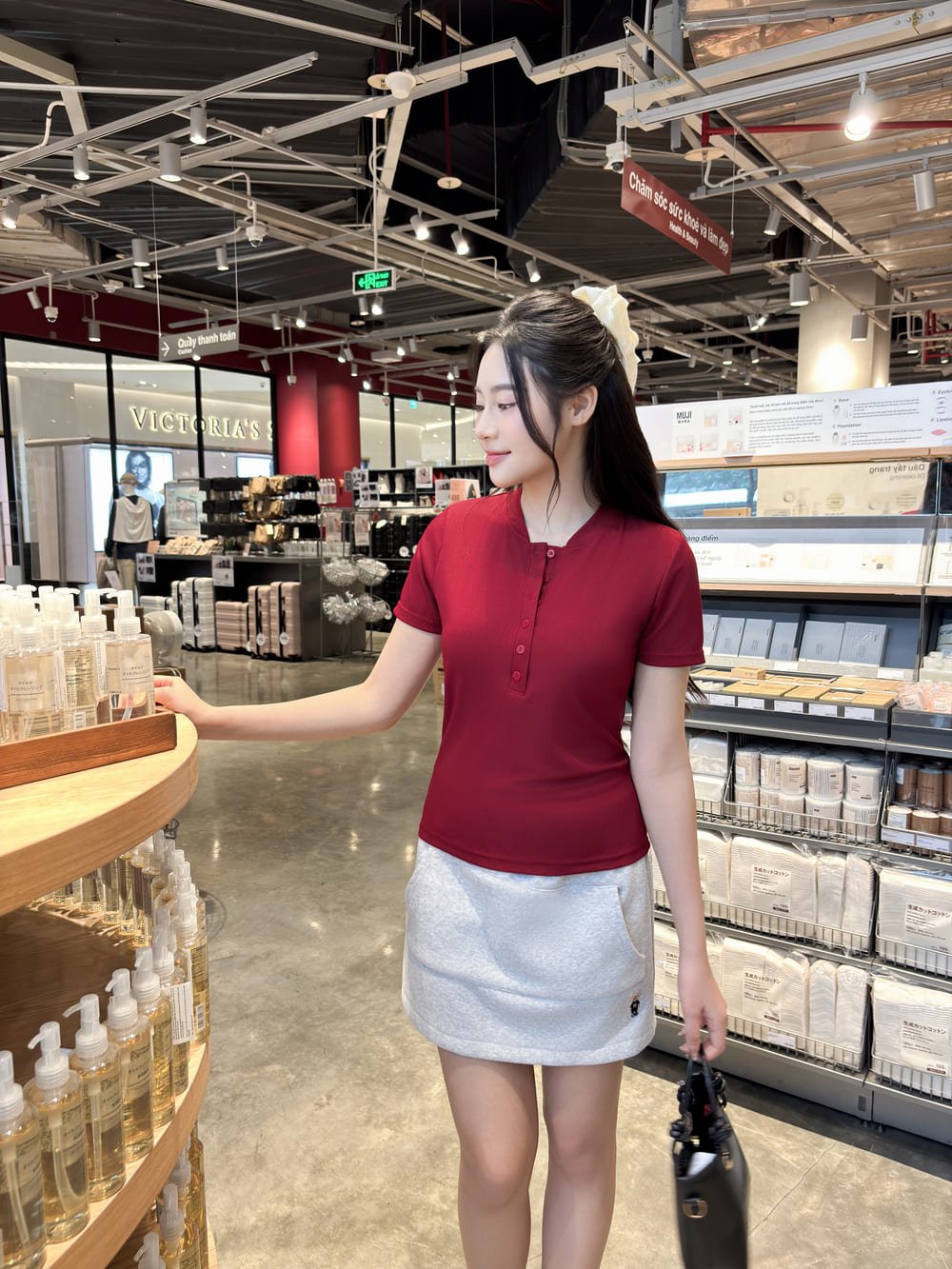 Người phụ nữ mặc áo đỏ, váy trắng, đứng trong cửa hàng mỹ phẩm, tay chạm vào kệ trưng bày sản phẩm.