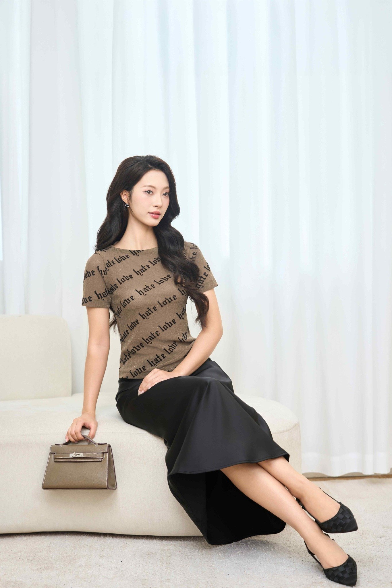 Người phụ nữ ngồi trên ghế, mặc áo in chữ, váy đen và cầm túi xách, nền là rèm trắng.