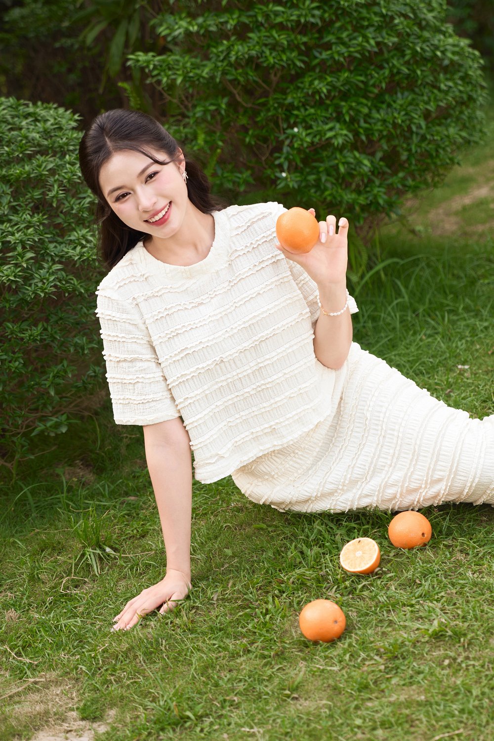 Người mặc trang phục trắng ngồi trên cỏ, cầm quả cam, xung quanh có nhiều trái cam khác.