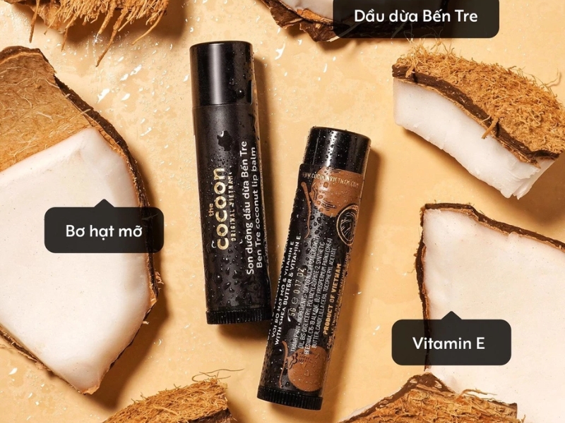 b01199b0-review-son-duong-moi-tot-10 Hai thỏi son dưỡng môi nằm trên bề mặt, xung quanh là các mảnh dừa, có chú thích thành phần tự nhiên.