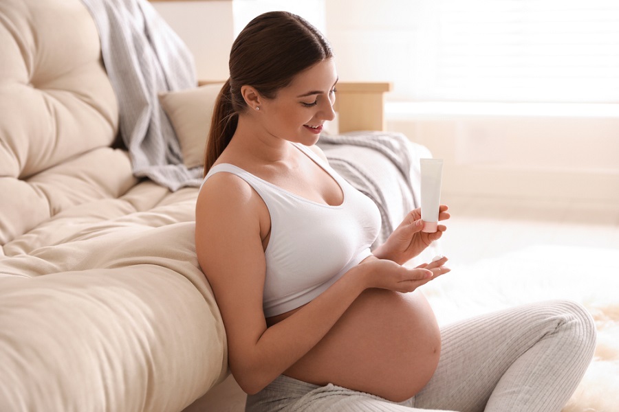 Người phụ nữ mang thai ngồi trên sàn, cầm tuýp kem dưỡng da, mỉm cười, trong không gian phòng khách sáng sủa.