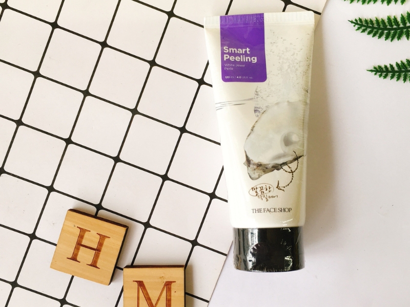 Tuýp mỹ phẩm trên nền gạch vuông trắng đen, cạnh hai khối gỗ có chữ "H" và "M", cùng cành cây xanh.