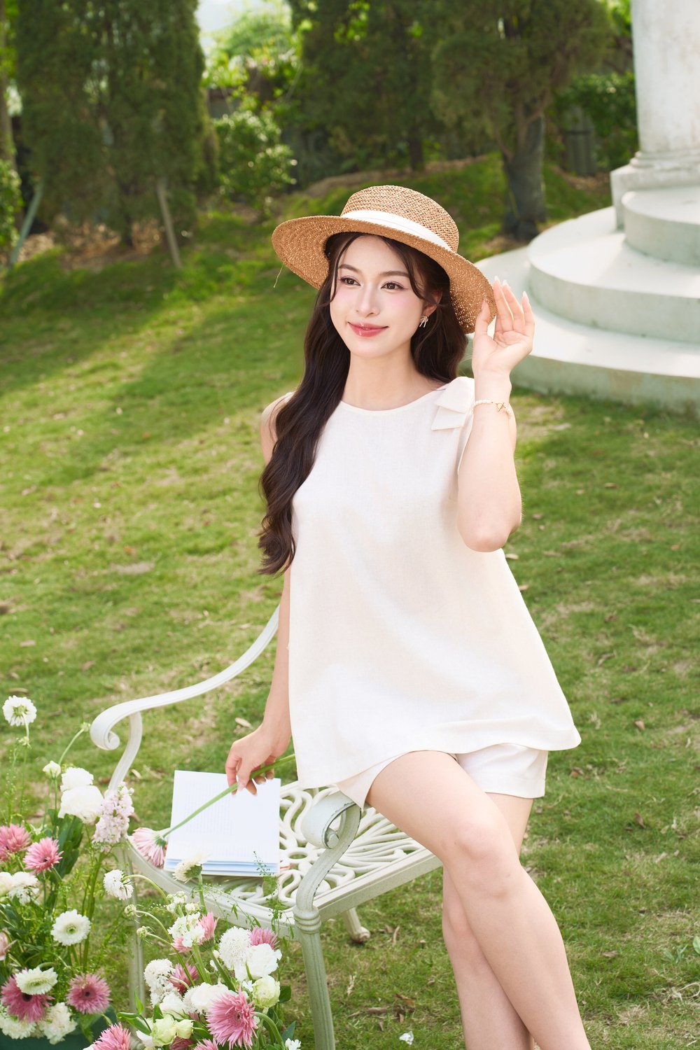 Người phụ nữ ngồi trên ghế trong vườn, đội nón và cầm sách, xung quanh có hoa tươi và cây xanh.
