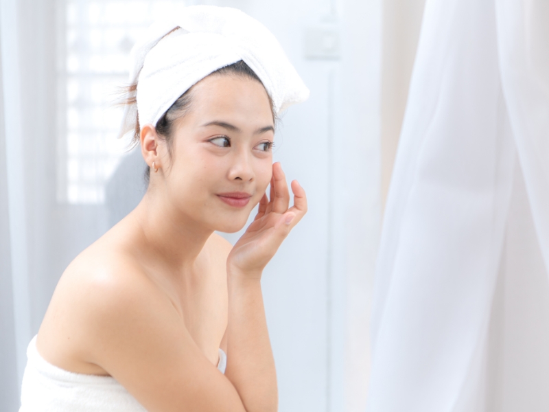 c3248b0a-review-mat-na-ngu-2 Người phụ nữ quấn khăn tắm, chạm nhẹ vào má, đứng trước gương trong phòng tắm sáng sủa.
