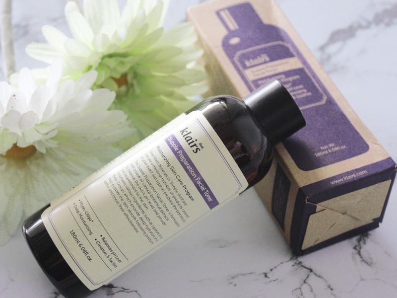 Chai toner nằm cạnh hộp giấy và hoa trắng trên nền đá cẩm thạch, nhãn hiệu có chữ "Klairs".