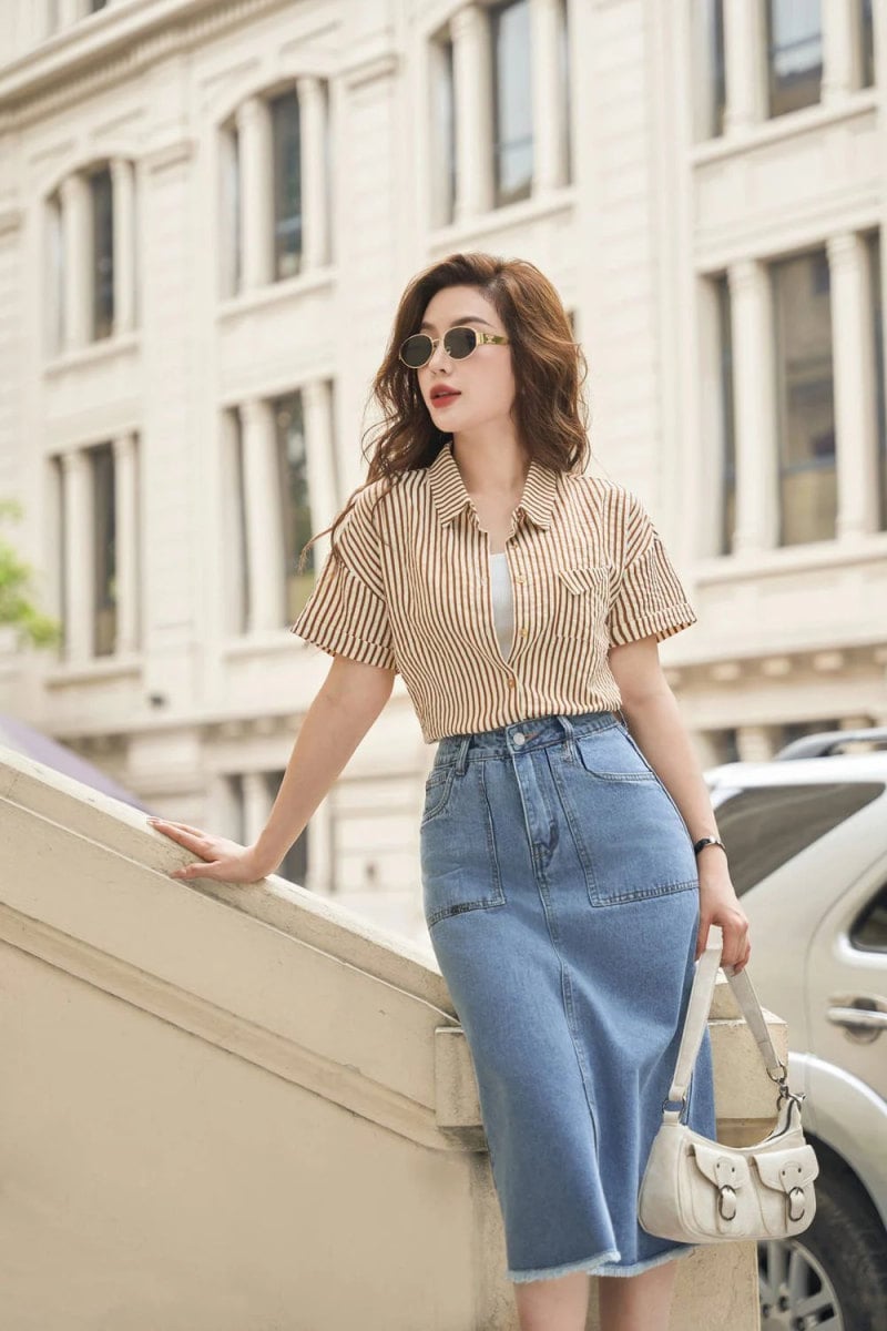 Người phụ nữ đeo kính râm, mặc áo sơ mi kẻ sọc và váy denim, đứng bên lan can ngoài trời.