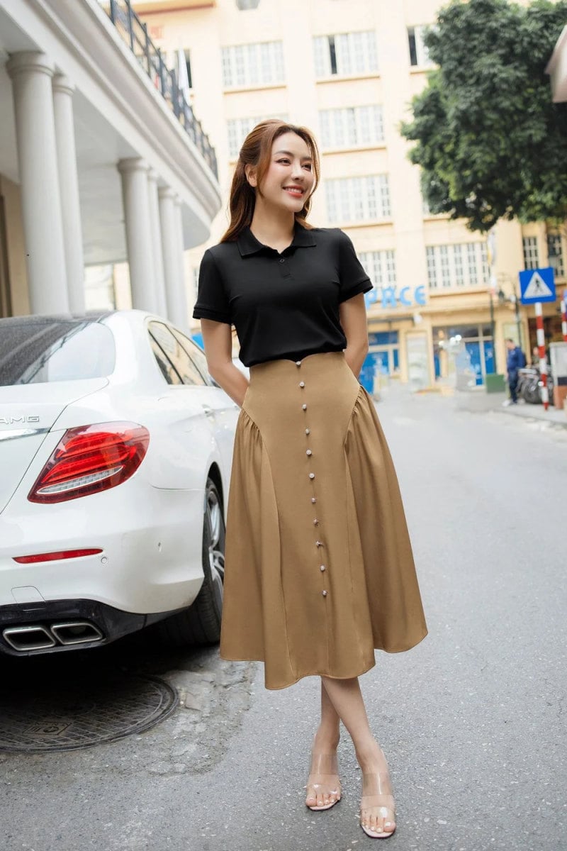 Người phụ nữ mặc áo đen, váy nâu đứng cạnh ô tô trắng trên con phố có tòa nhà phía sau.