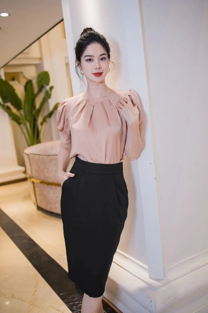 Người phụ nữ mặc áo hồng và váy đen, đứng tựa vào tường trong không gian nội thất sang trọng.
