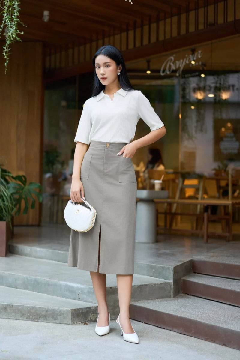 Người phụ nữ mặc trang phục thanh lịch đứng trước cửa hàng, tay cầm túi xách nhỏ, nền gỗ và cây xanh.