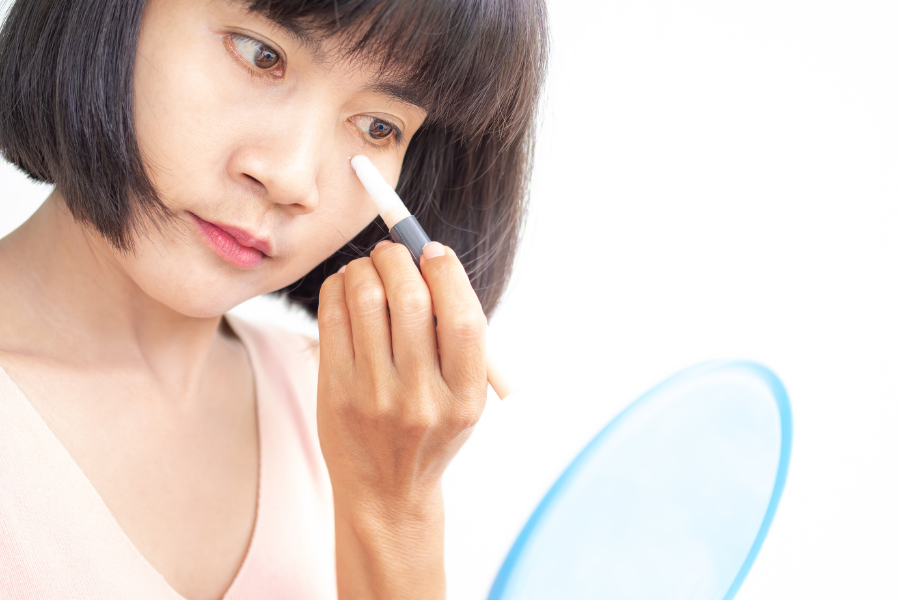 Người phụ nữ đang sử dụng bút trang điểm gần mắt, cầm gương tròn màu xanh dương.