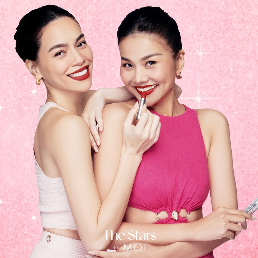 Hai người phụ nữ cười tươi, một người cầm son, nền hồng lấp lánh.