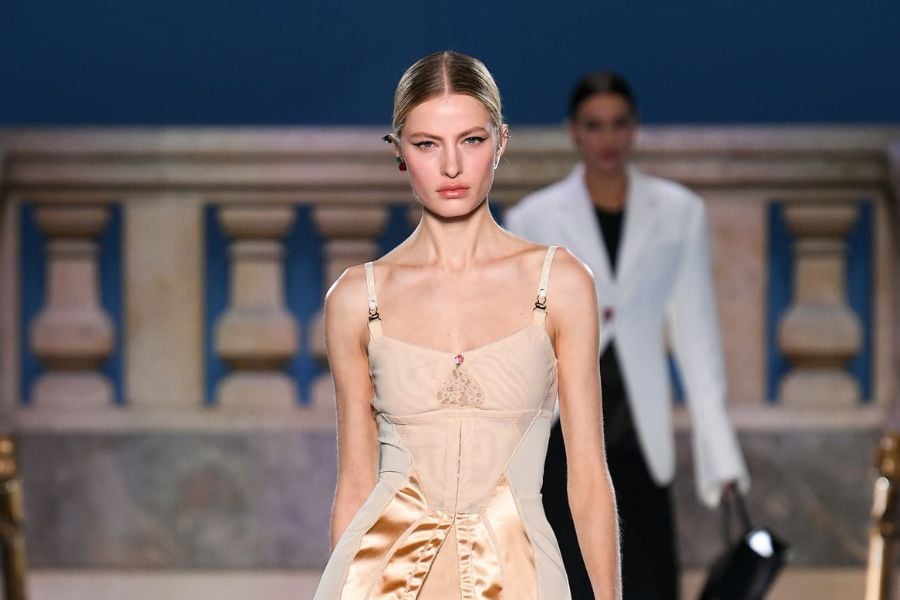 Người mẫu trình diễn trang phục trên sàn catwalk, nền có người khác mặc vest trắng.