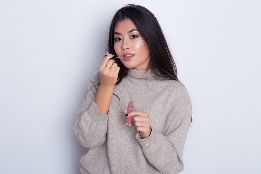 Người phụ nữ mặc áo len màu be, đang cầm son và chuẩn bị thoa lên môi, nền trắng.