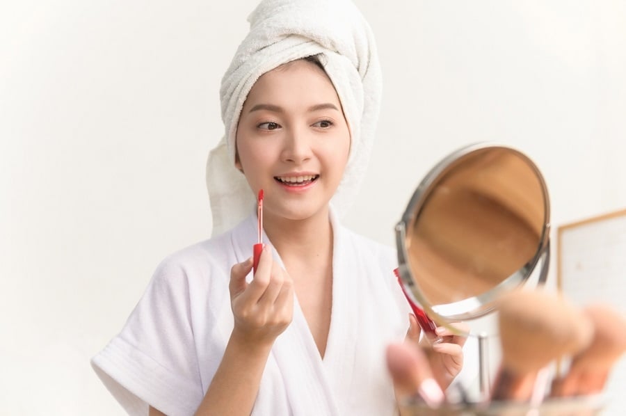Người cầm gương trang điểm, quấn khăn trắng trên đầu, chuẩn bị thoa son môi đỏ, xung quanh có cọ trang điểm.