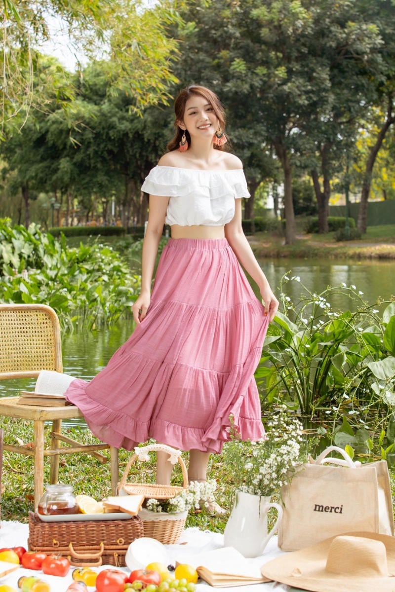 Người phụ nữ mặc áo trắng và váy hồng đứng bên bàn picnic ngoài trời, xung quanh là cây cối xanh tươi.