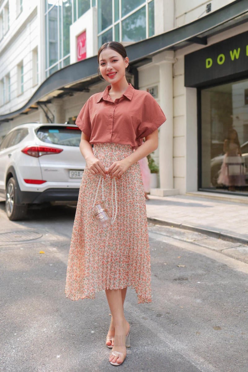 Người phụ nữ đứng trên phố, mặc áo sơ mi nâu và váy hoa, cầm túi xách nhỏ, phía sau là xe ô tô trắng.