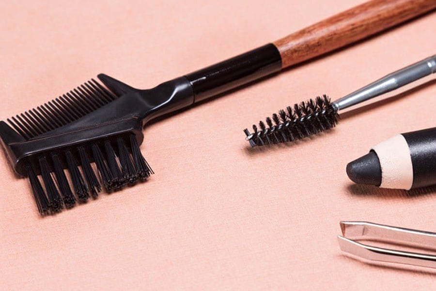 Bộ dụng cụ trang điểm gồm cọ, chổi mascara, bút kẻ mắt và nhíp trên nền vải màu hồng nhạt.